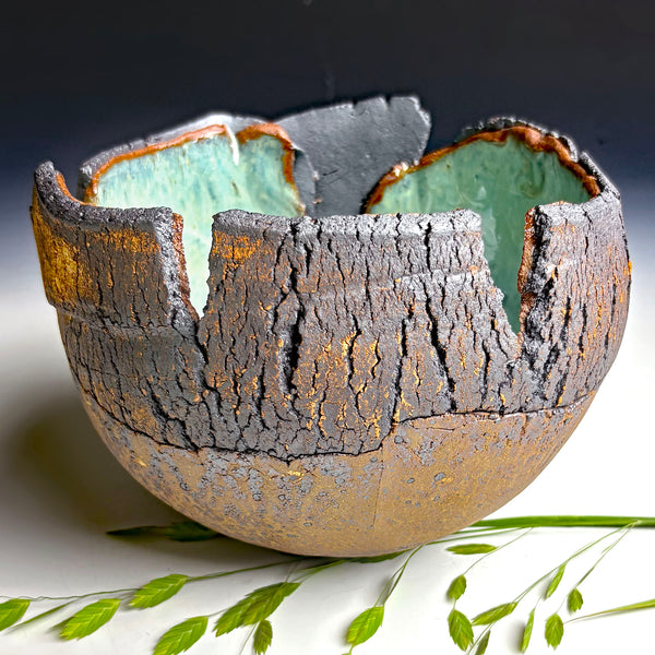 A handmade ceramic bowl with a textured, bark-like exterior in dark gray and gold, and a glossy light green interior, styled with green leaves on a white surface against a dark background.