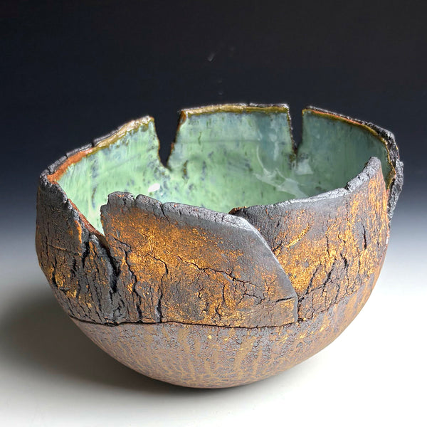 A handmade ceramic bowl with a unique cracked, leaf-like exterior in dark brown and gold tones, and a smooth, light green glazed interior, shown on a white surface against a dark background.