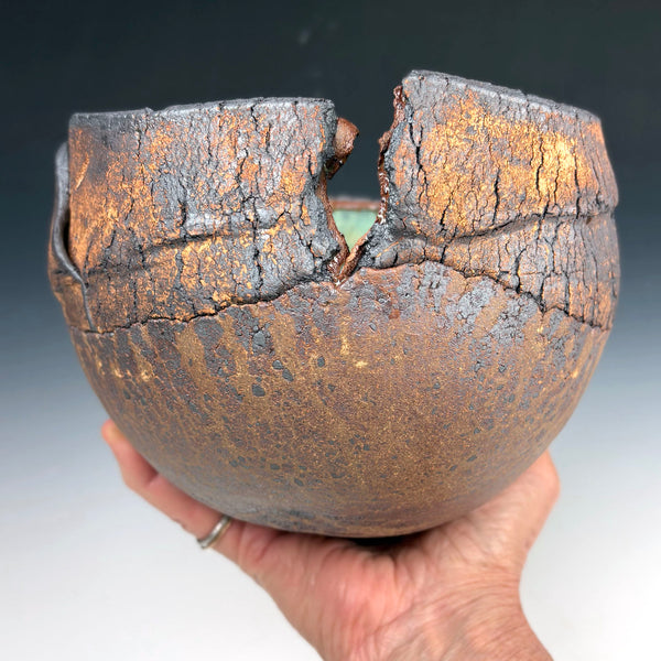 A handmade ceramic bowl with a heavily cracked, textured surface in earthy brown, black, and gold tones, held in a person's hand against a dark gradient background.