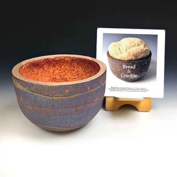 A handmade ceramic bowl with a textured orange-brown glazed interior and a blue-gray glazed exterior with gold crackle detailing, displayed next to a small book titled 'Bread & Crucible' on a wooden stand against a dark studio background.