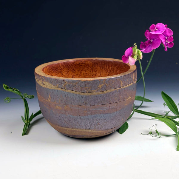 A handmade wooden bowl with a textured, crackled finish in earthy brown and muted purple-gray tones, styled with green leaves and pink flowers against a dark blue gradient background.