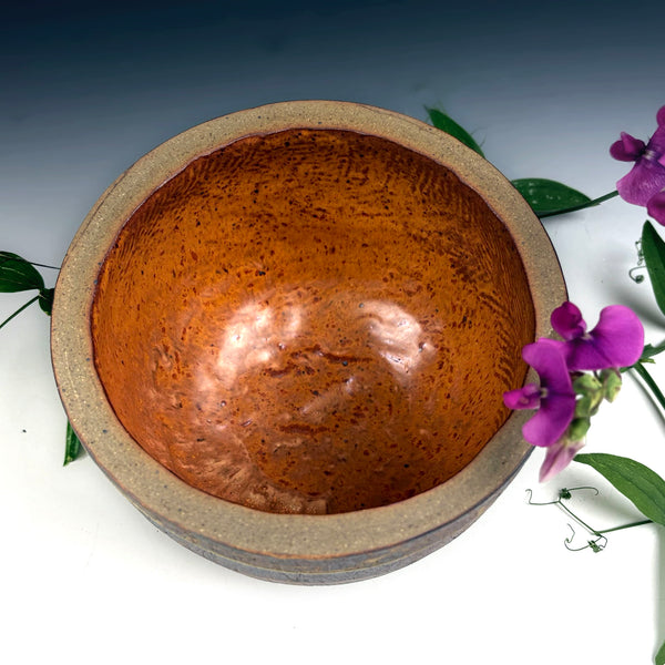 A handmade ceramic bowl with a textured, speckled brown interior and a matte beige-gray exterior, styled with purple flowers and green leaves on a white surface against a dark gradient background.