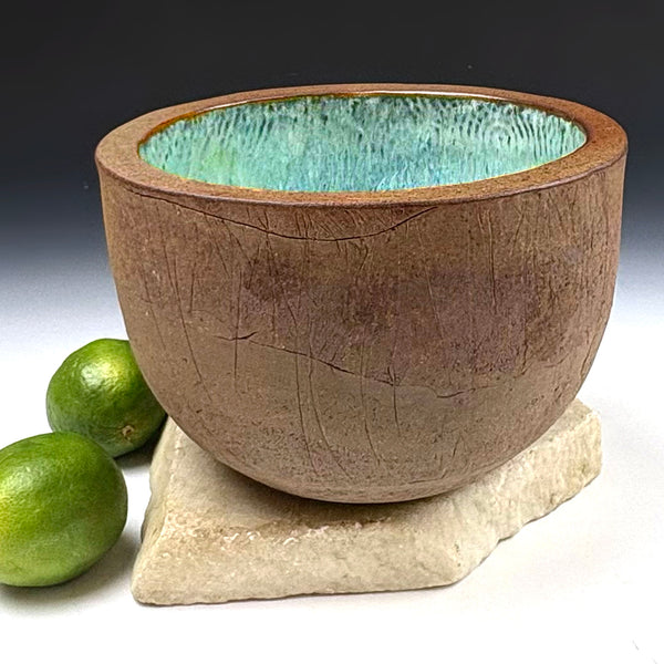 A handmade ceramic bowl with a textured brown exterior and a glossy turquoise-blue interior, displayed on a stone base with two limes in the background.