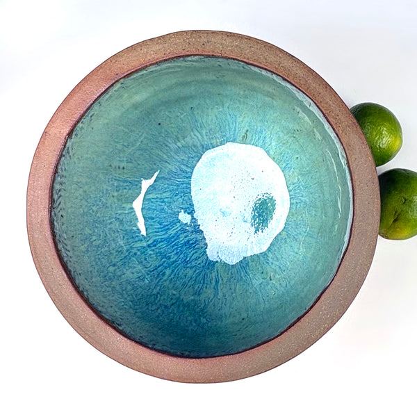 A round handmade ceramic bowl with a textured brown clay rim and a blue-green glazed interior featuring a white splatter design, shown on a white background with two limes as props.