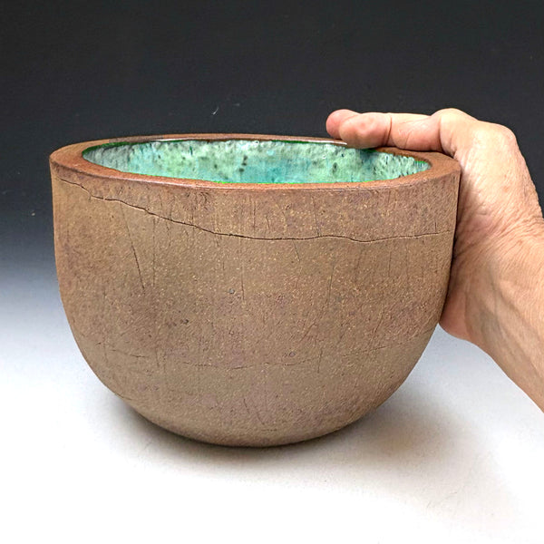 A handmade ceramic bowl with a textured brown exterior and a green glazed interior, held by a hand against a dark gray background.