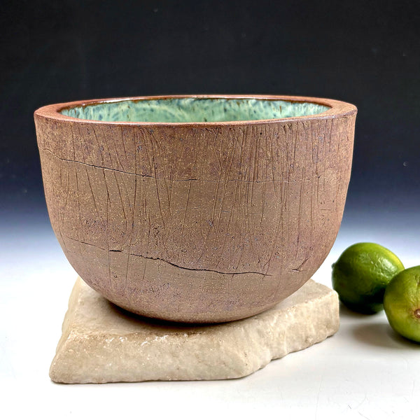 A handmade ceramic planter with a textured, earthy brown exterior and a green glazed interior, displayed on a light stone slab with two limes in the background against a dark gradient backdrop.