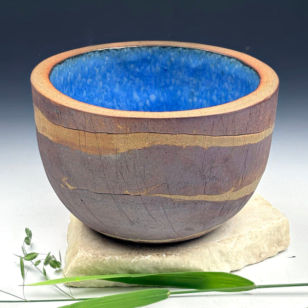 A handmade ceramic bowl with a textured, earthy brown exterior featuring horizontal gold-toned bands, and a vibrant blue glazed interior, displayed on a stone slab with green leaves.