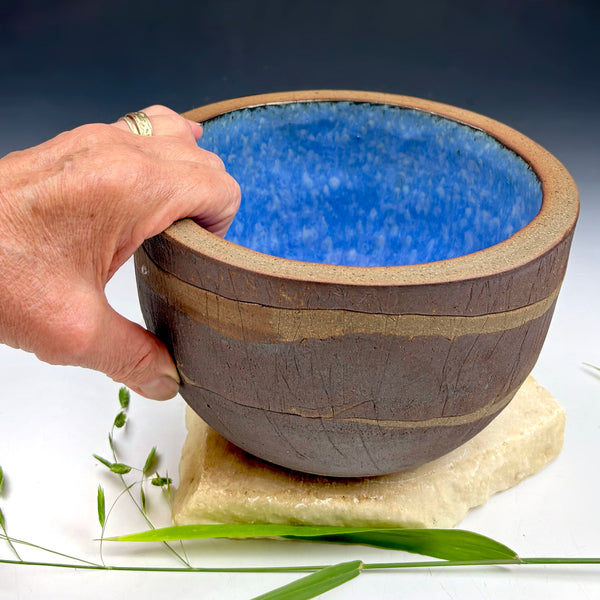 A handmade ceramic bowl with a textured blue interior and a dark brown, earthy exterior, held by a hand resting on a stone slab with green grass in the foreground.