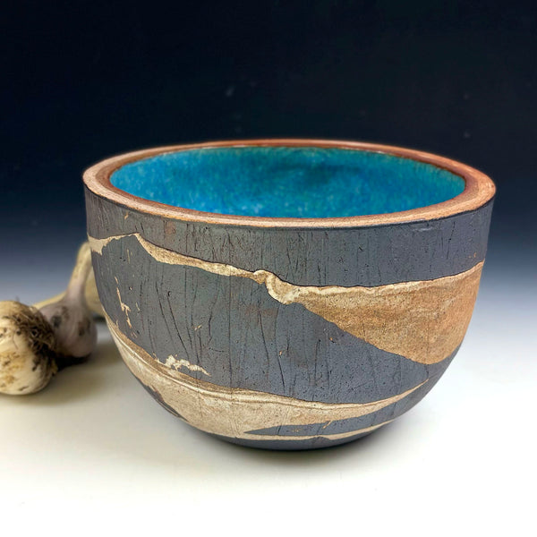 A handmade ceramic bowl with a textured dark gray exterior, natural beige and brown clay inlay patterns, and a vibrant turquoise blue glazed interior, shown on a white surface with a dark gradient background.