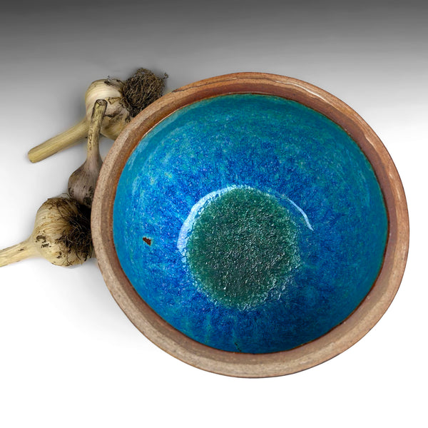 A handmade ceramic bowl with a vibrant blue and green glazed interior and a natural brown clay rim, shown on a white background with garlic bulbs as styling props.