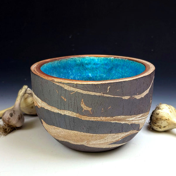 A handmade ceramic bowl with a dark gray exterior featuring horizontal beige wood-grain patterns and a vibrant blue glazed interior, styled with garlic bulbs on a white surface against a dark background.