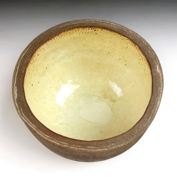 A handmade ceramic bowl with a textured, rustic brown exterior and a glossy, pale yellow-green glazed interior.