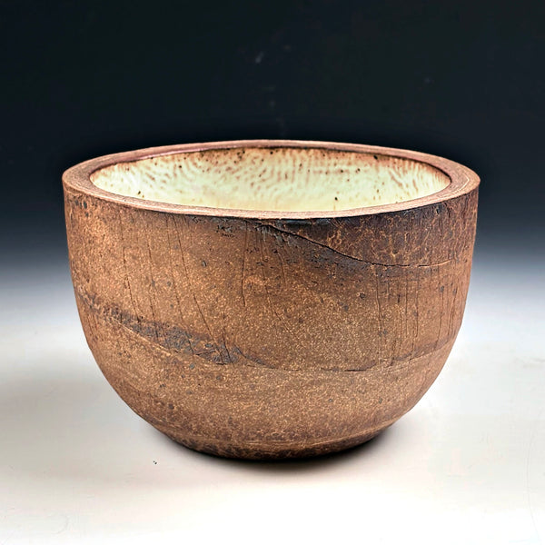 A handmade ceramic bowl with a rustic, textured brown exterior and a contrasting light beige speckled glaze interior, set against a dark gradient background.