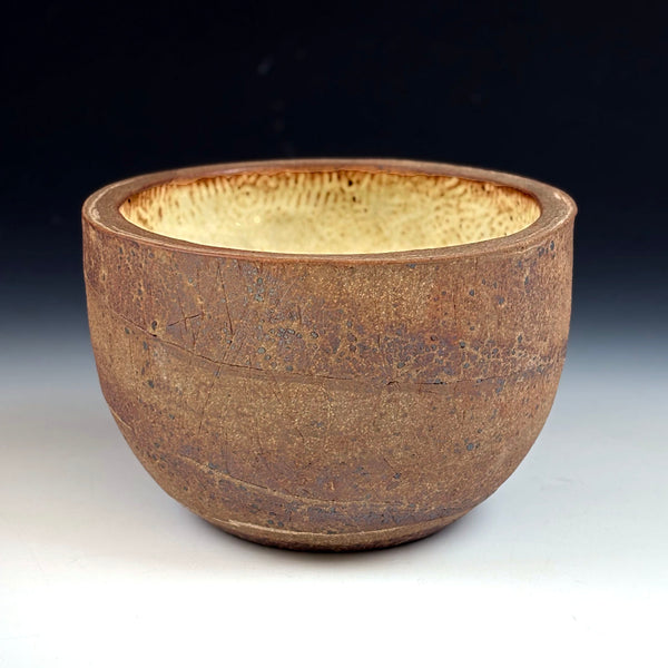 A handmade ceramic bowl with a rustic, textured brown exterior and a contrasting light beige, speckled interior, set against a dark-to-light gradient background.
