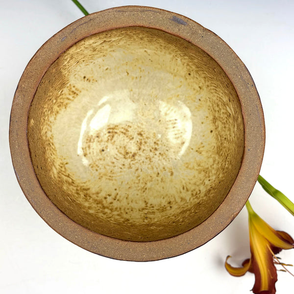 A handmade ceramic bowl with a speckled beige and brown interior glaze and a textured brown exterior, photographed from a top-down angle on a white background with a yellow flower stem as a prop.