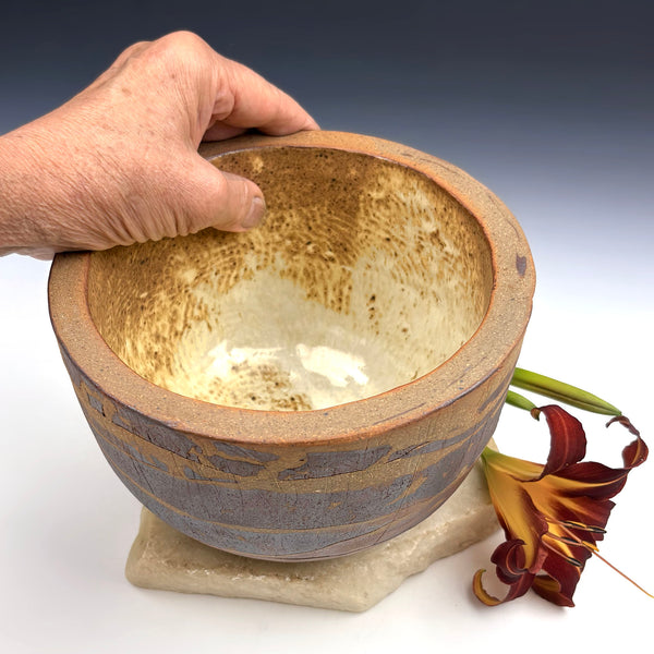 A handmade ceramic bowl with a rustic speckled brown interior and a textured gray-blue exterior, held by a hand to show its depth. The bowl sits on a small stone base next to a dried marigold flower against a gradient gray background.