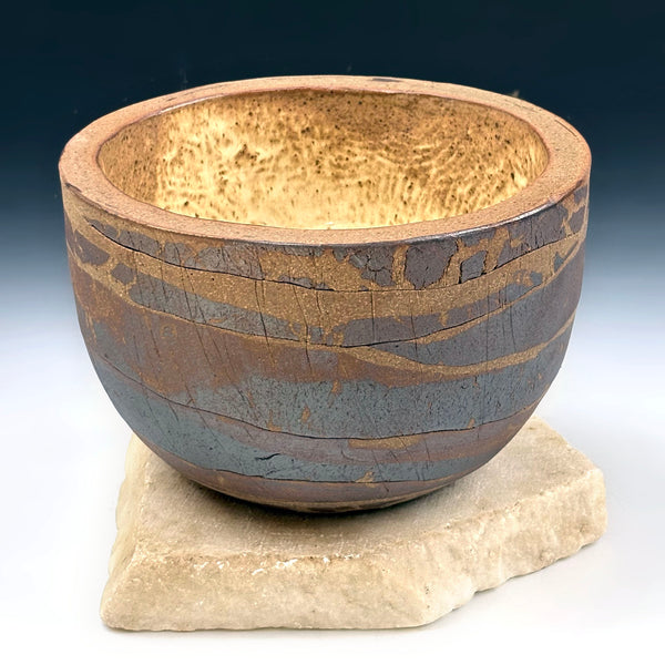 A handmade ceramic bowl with a textured, hammered finish and horizontal metallic gold and bronze bands, displayed on a rustic stone slab against a dark blue gradient background.