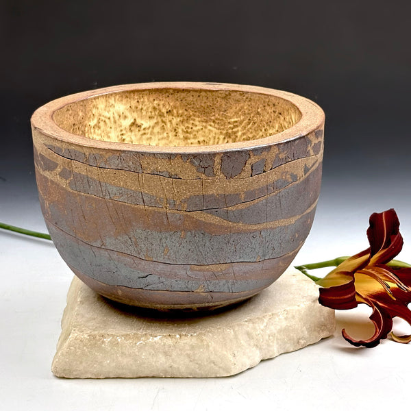 A handmade ceramic bowl with a textured, crackled glaze in earthy brown, beige, and gray tones, displayed on a small stone pedestal with a dark flower prop against a dark gray background.
