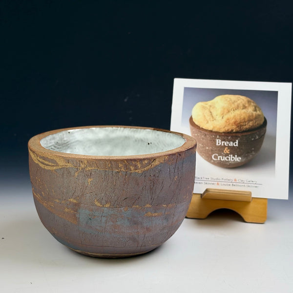 A handmade ceramic bread bowl with a rustic brown and gold glaze sits on a white surface against a dark background, with a printed card showing a baked loaf behind it on a small wooden stand.