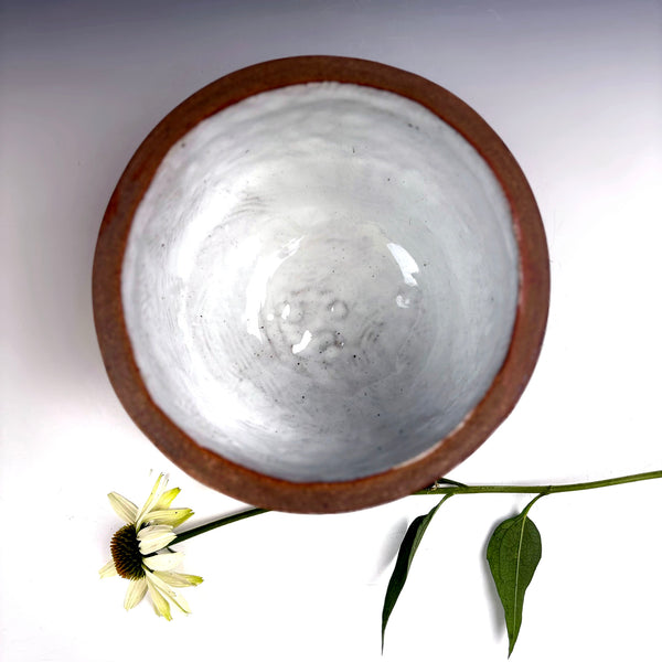 A handmade ceramic bowl with a rustic brown rim and a glossy, textured off-white interior, photographed from a top-down angle on a white background with a small white flower and green stem as props.