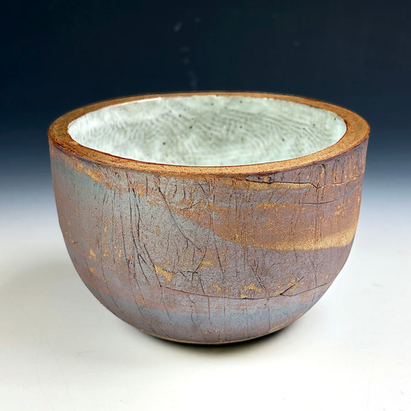 A handmade ceramic bowl with a textured, earthy exterior in brown and gray tones, and a contrasting light gray glazed interior, shown against a dark-to-light gradient background.