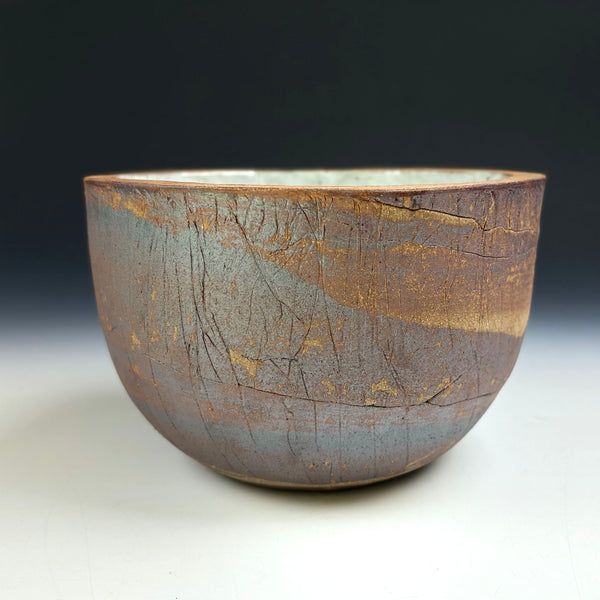 A handmade ceramic bowl with a textured, earthy glaze featuring brown, gray, and gold tones, photographed against a dark-to-light gradient studio background.
