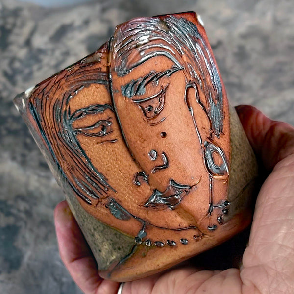 A handmade ceramic pot with an embossed stylized face design, held in a person's hand against a textured gray background.