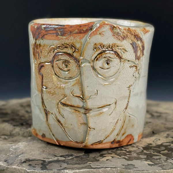 A handmade ceramic cup featuring a stylized face design with brown and orange glaze accents on a light gray base, set on a textured stone surface against a dark background.