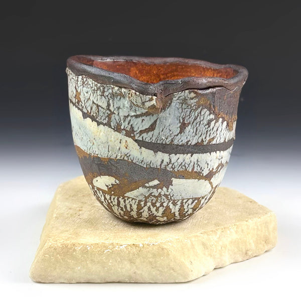 A handmade ceramic cup with a rustic, irregular rim and a textured glaze featuring brown, white, and gray patterns, displayed on a light beige stone block against a dark gray gradient background.
