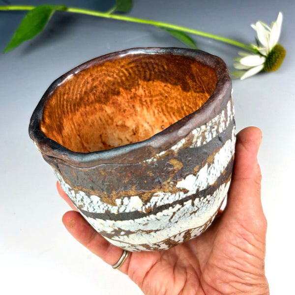 A handmade ceramic cup with a rustic, hand-painted finish, held in a person's hand. The cup features a textured, hammered metallic bronze-brown interior and a patterned exterior with white, dark brown, and black glazes.