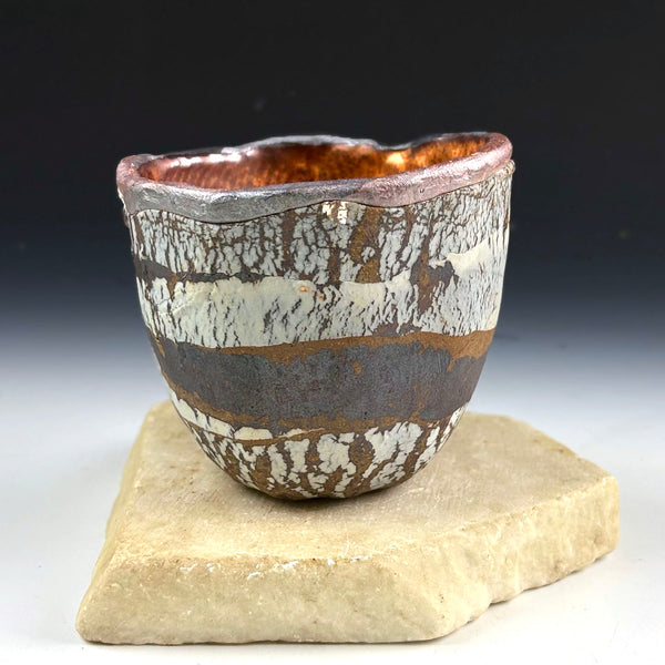 A handmade ceramic cup with a textured, crackled glaze in earthy tones of brown, gray, and off-white, sitting on a natural stone slab against a dark gradient background.