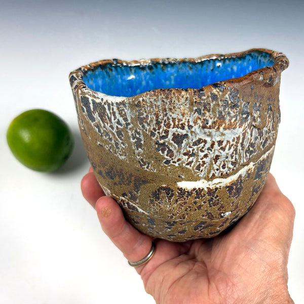 A handmade ceramic bowl with a rustic, crackled brown and gold exterior and a vibrant blue glazed interior, held in a person's hand with a lime in the background.