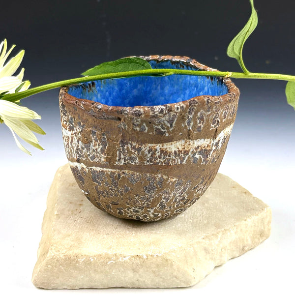 A handmade ceramic bowl with a textured brown and white glaze and a vibrant blue interior, displayed on a light stone slab with a white flower stem resting across the rim.
