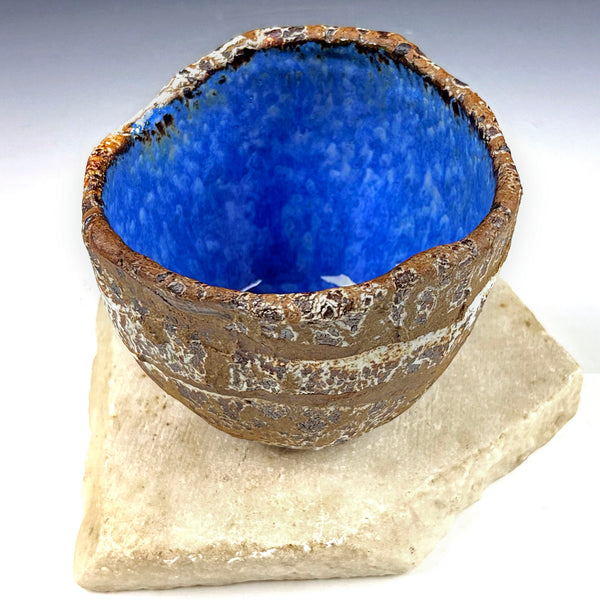 A handmade ceramic bowl with a vibrant blue glazed interior and a textured, earthy brown exterior, displayed on a light beige stone block against a white background.
