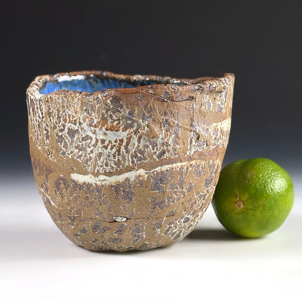 A handmade ceramic bowl with a textured, crackled brown and beige glaze and a visible blue interior, shown next to a green lime on a white surface against a dark gray background.