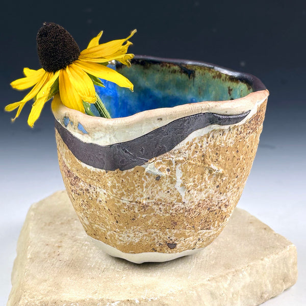 A handmade ceramic cup with a rustic, textured beige exterior and a glossy blue interior, decorated with a dark brown band near the rim. A yellow flower is tucked into the side of the cup, which is displayed on a light-colored stone block against a dark gradient background.