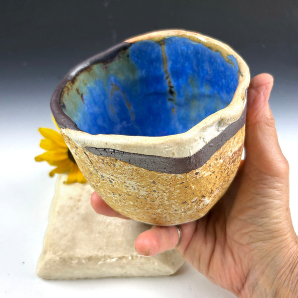 A handmade ceramic bowl with a textured beige exterior and a vibrant blue glazed interior, held by a hand over a small stone pedestal.