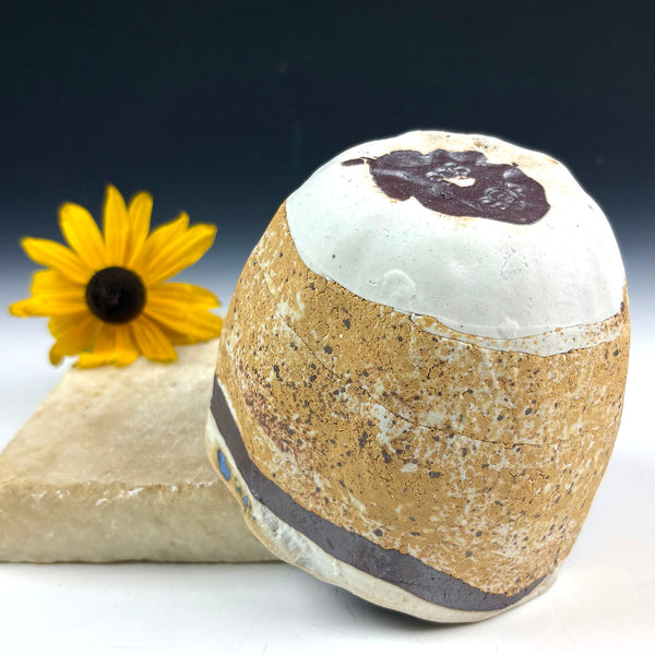 A rounded, textured stone or ceramic object with a white top section, a tan speckled middle section, and dark brown bands, displayed on a white surface with a yellow flower and stone prop against a dark gradient background.