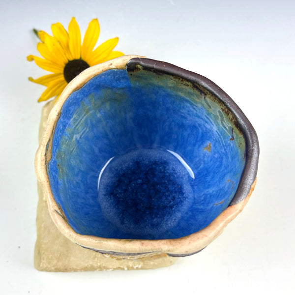 A handmade ceramic cup with a vibrant blue glazed interior and a rustic, unglazed beige rim, shown on a white background with a yellow flower prop.