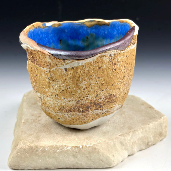 A handmade ceramic cup with a rough, textured tan exterior and a glossy blue interior, displayed on a flat stone slab against a dark gradient background.