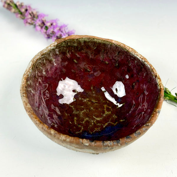A handmade ceramic bowl with a glossy deep red and purple glaze, showing a rustic brown clay edge, photographed on a white background with a sprig of purple flowers in the background.
