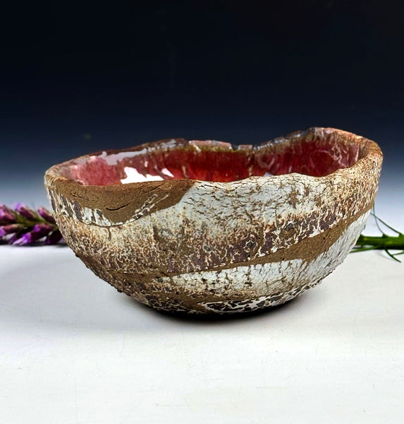 A handmade ceramic bowl with a textured, earthy brown and white exterior and a glossy red interior, displayed on a white surface with a dark background.