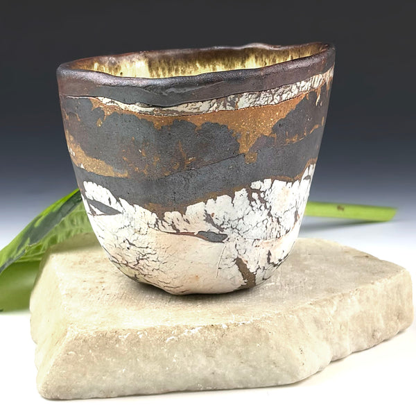 A handmade ceramic cup with a textured, crackled glaze in dark brown, black, white, and gold tones, displayed on a rustic stone block with green leaves in the background.