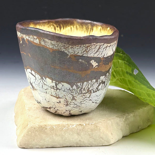 A handmade ceramic cup with a textured, crackled glaze in white, brown, and gold tones, displayed on a light-colored stone slab with a green leaf against a dark gradient background.