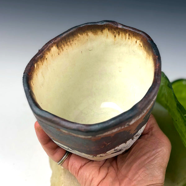 A handmade ceramic cup with a rustic dark brown exterior and a glossy cream-colored interior, held in a person's hand against a neutral gray background.