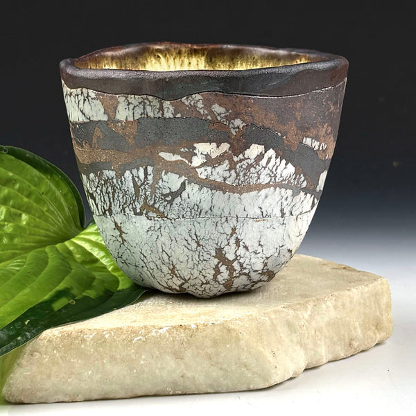 A handmade ceramic cup with a textured, crackled glaze in brown, white, and black, resting on a light stone slab with a green leaf prop against a dark background.