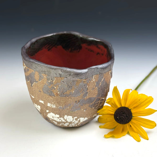 A handmade ceramic cup with a textured, crackled metallic finish in silver, gold, and white, featuring a glossy dark red interior. A yellow daisy flower is placed next to it on a white surface with a dark gray gradient background.