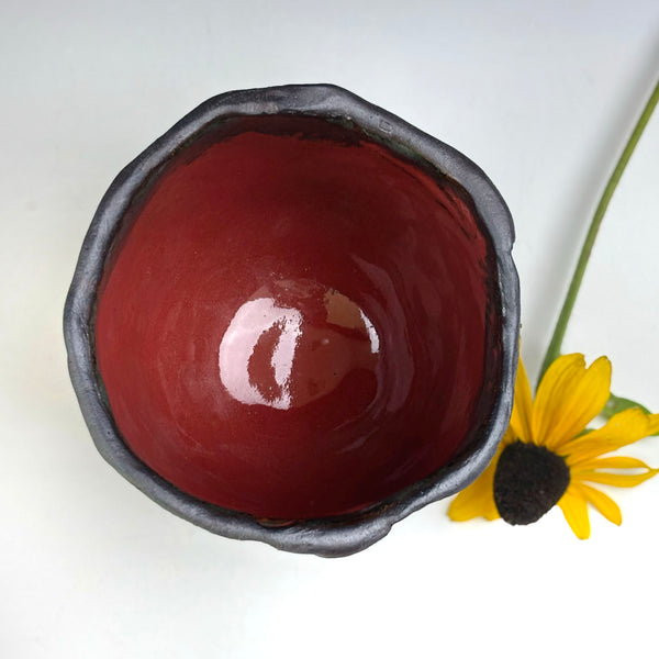 A handmade ceramic bowl with a glossy dark red interior and a textured, dark charcoal gray exterior, shown from a top-down view next to a yellow flower on a white background.