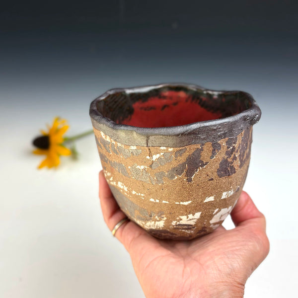 A handmade ceramic cup with a rustic, textured glaze in earthy brown, beige, and black, held by a hand. The interior of the cup is a deep terracotta red.