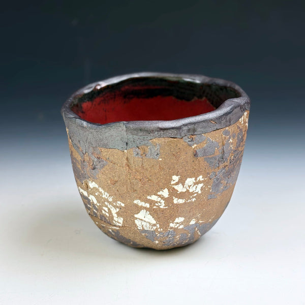 A handmade ceramic cup with a textured, metallic gold and silver exterior and a glossy dark red interior, set against a dark gray gradient background.
